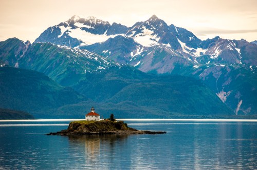 A small lighthouse on an island surrounded by calm waters, with snow-capped mountains in the background.