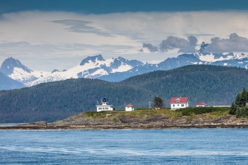 A scenic view of a lighthouse and houses on a rocky shore, with snow-capped mountains in the background and a calm sea.
