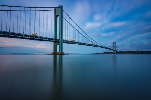 A long exposure shot of a suspension bridge over calm water, with clouds moving in the blue sky above.