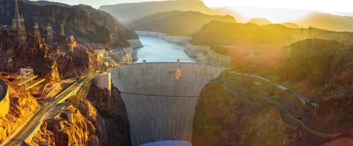 A panoramic view of a large dam surrounded by mountains, with a river flowing below and sunlight illuminating the scene.