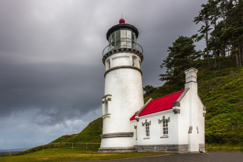 A white lighthouse with a red roof stands on a grassy hill under a cloudy sky.
