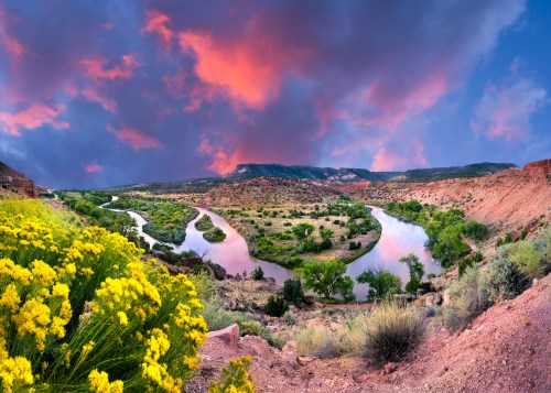 A scenic river winding through lush greenery, framed by vibrant yellow flowers and a dramatic sunset sky.