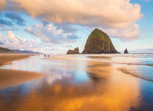 A scenic beach at sunset with a rocky outcrop, reflecting colorful clouds and water, and people walking along the shore.