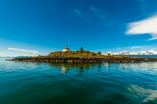 A serene coastal scene featuring a lighthouse on an island, reflecting in calm waters under a clear blue sky.