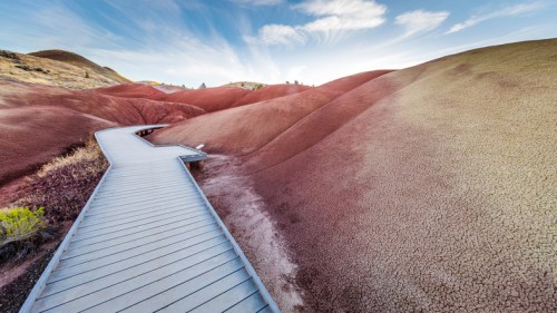 A wooden walkway winds through colorful, undulating hills under a blue sky with wispy clouds.