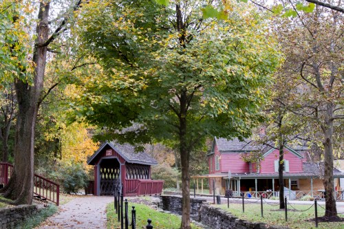 A scenic path leads to a red covered bridge, surrounded by trees and a historic house in autumn foliage.