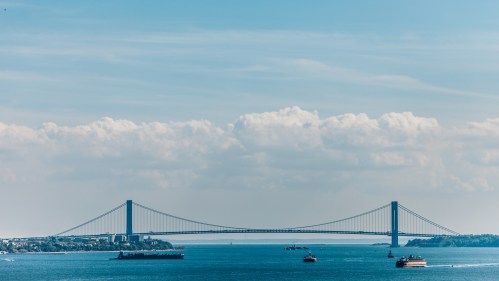 A suspension bridge spans a calm waterway, with ships passing beneath and fluffy clouds in a blue sky above.