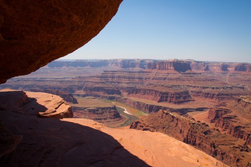 A panoramic view of a canyon landscape with red rock formations and a winding river below, under a clear blue sky.