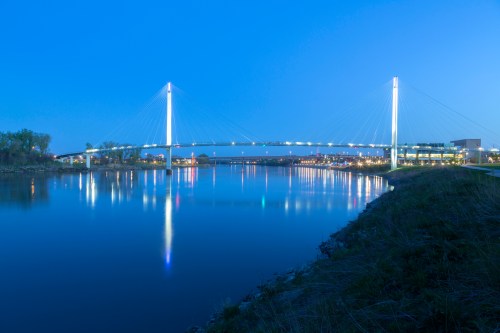 A modern cable-stayed bridge illuminated at dusk, reflecting in a calm river with city lights in the background.