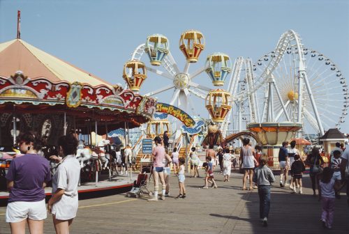 A vibrant amusement park scene with a carousel, Ferris wheel, and crowds enjoying a sunny day on the boardwalk.