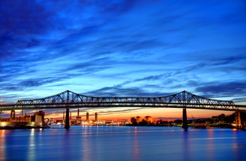 A bridge spans a river at sunset, with vibrant blue and orange skies reflecting on the water.