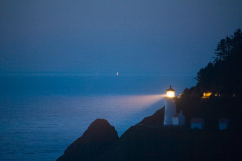 A lighthouse beams light over a calm ocean at dusk, with distant lights visible on the horizon.