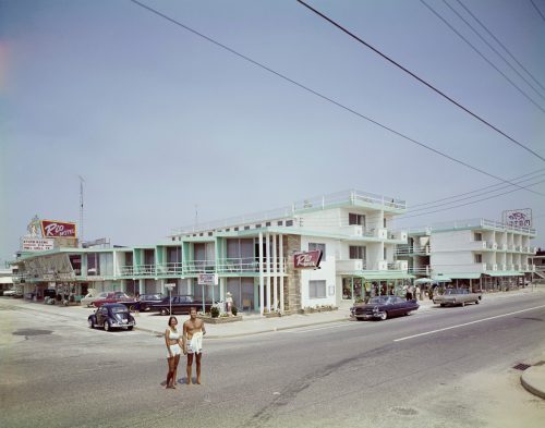 Two people stand on a street in front of a retro motel, with classic cars parked nearby and shops in the background.