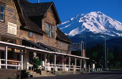 Historic wooden buildings with a café sign, set against a backdrop of a snow-capped mountain under a clear blue sky.