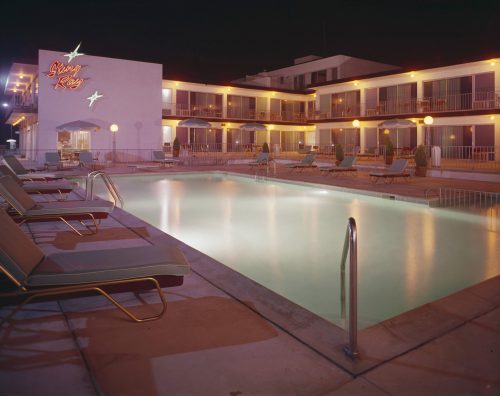 A nighttime view of a motel pool area, featuring lounge chairs and illuminated buildings with a retro sign.