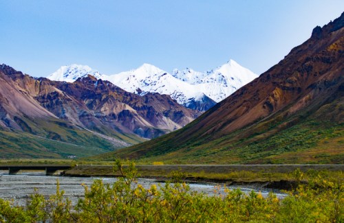 Snow-capped mountains rise above colorful, rugged terrain with a river and greenery in the foreground.