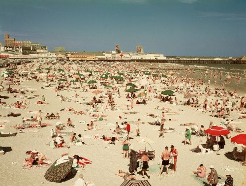 A crowded beach scene with numerous umbrellas, sunbathers, and a pier in the background under a clear blue sky.