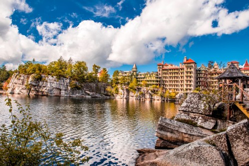 A scenic view of a lake surrounded by rocky cliffs and a historic building under a blue sky with fluffy clouds.