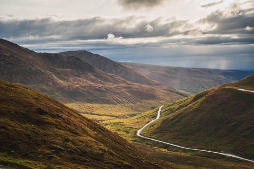 A winding road cuts through a lush valley surrounded by mountains under a cloudy sky.