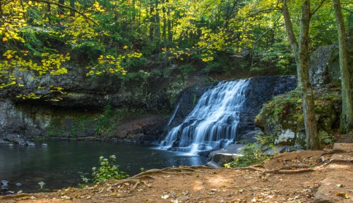 A serene waterfall cascades into a calm pool, surrounded by lush green trees and autumn leaves.