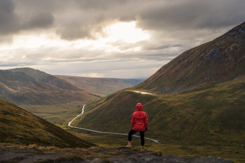 A person in a red jacket stands on a hillside, overlooking a valley with winding roads and dramatic clouds above.