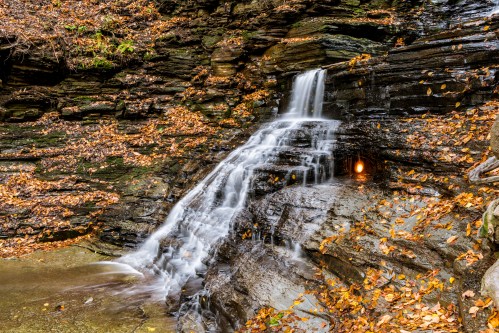 A serene waterfall cascades over rocky terrain, surrounded by autumn leaves and a tranquil pool below.