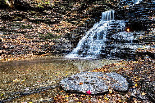A serene waterfall cascades over rocky layers, surrounded by autumn leaves and a calm pool below.
