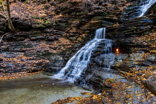 A serene waterfall cascades over rocky layers, surrounded by autumn leaves and a tranquil pool below.