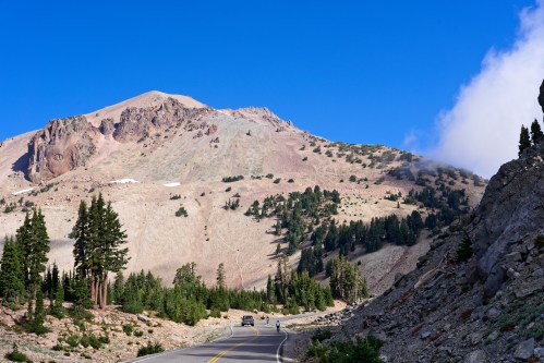 A winding road leads through a mountainous landscape with trees, under a clear blue sky.