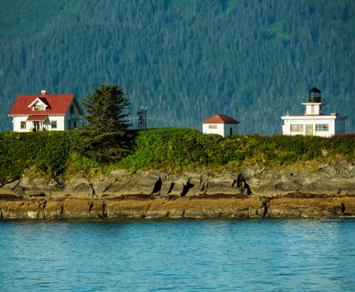 A coastal scene featuring a house and a lighthouse on a rocky shore, surrounded by lush green hills and calm water.