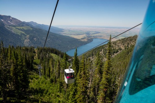 A scenic view from a gondola overlooking a lake and mountains, surrounded by lush green trees.