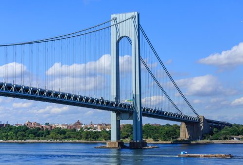 A large suspension bridge spans a river under a blue sky with fluffy clouds, with greenery and buildings in the background.
