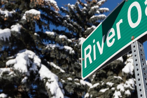 Green street sign reading "River Oak" with snow-covered trees in the background under a clear blue sky.