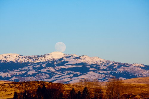 A full moon rises over snow-capped mountains under a clear blue sky.