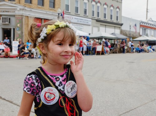 A smiling girl in a floral headband and colorful outfit waves at a parade, with people and tents in the background.