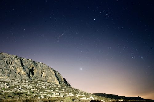 A starry night sky over a rocky landscape, with a bright planet and a meteor streaking across the scene.