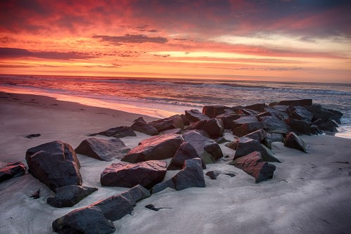 Sunset over a beach with rocky formations, casting vibrant colors across the sky and reflecting on the water.