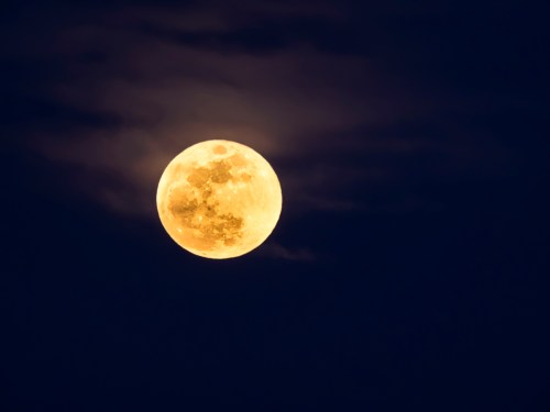 A bright full moon glowing in a dark night sky, partially surrounded by wispy clouds.