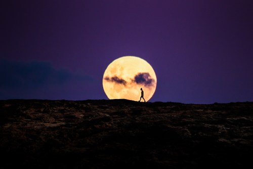 A silhouette of a person walking on a rocky landscape with a large, glowing full moon in the background.
