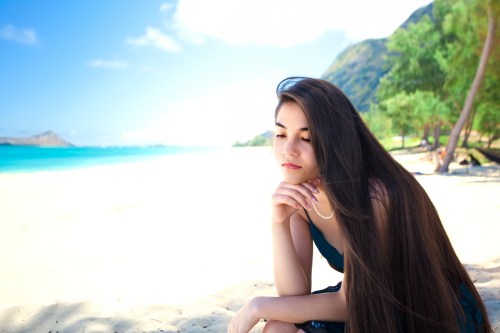 A young woman with long hair sits thoughtfully on a beach, with turquoise water and green trees in the background.