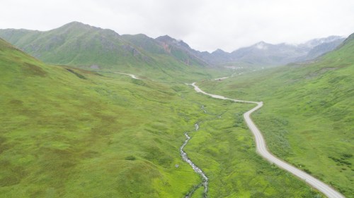Aerial view of a winding road through lush green hills and a small stream in a mountainous landscape under cloudy skies.