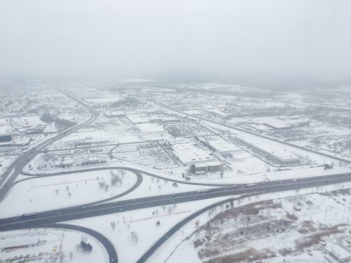Aerial view of a snowy landscape with roads, parking lots, and buildings partially obscured by fog.