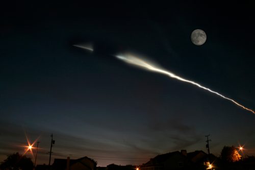A night sky with a bright moon and a streak of light, possibly from a rocket launch, above silhouetted houses.