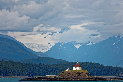A lighthouse on a rocky island surrounded by mountains and a cloudy sky, with a glacier in the background.