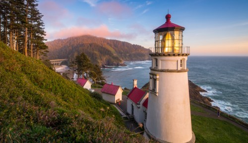 A scenic lighthouse on a cliff overlooking the ocean, surrounded by trees and mountains at sunset.