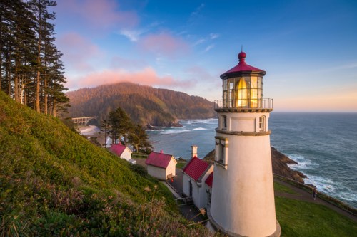 A scenic lighthouse on a cliff overlooking the ocean, surrounded by trees and mountains at sunset.