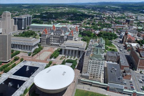 Aerial view of a cityscape featuring modern and historic buildings, green spaces, and distant mountains under a clear sky.