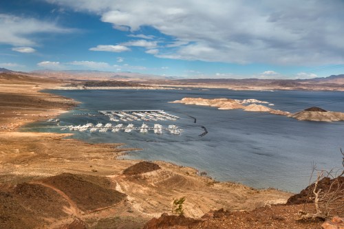 A scenic view of a marina on a lake, surrounded by arid land and distant mountains under a partly cloudy sky.