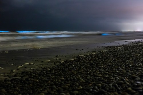 A dark beach scene at night with waves gently lapping at a pebbly shore under a cloudy sky.