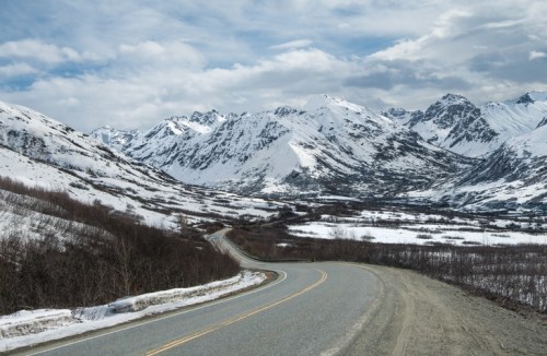A winding road leads through snow-covered mountains under a cloudy sky.